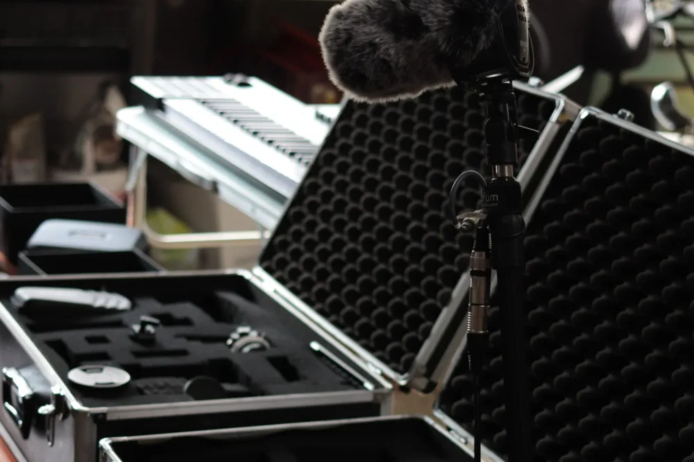 Professionelles Tonaufnahmemikrofon mit Fellwindschutz, neben geöffneten Transportkoffern für Filmausrüstung und einem Keyboard im Hintergrund.