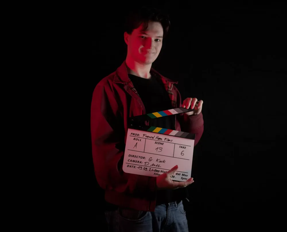 Porträt von Gerrit Kock von funeralhomefilms, der eine Filmklappe vor einer schwarzen Kulisse hält und lächelt. Er trägt eine rote Jacke und einen schwarzen Pullover.